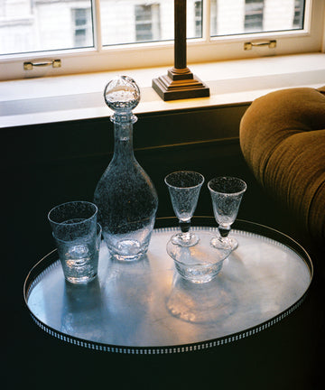 Two Lauren Wine Glasses are arranged on a round tray alongside a glass decanter, a tumbler, and a small bowl in front of a window showcasing a city view.