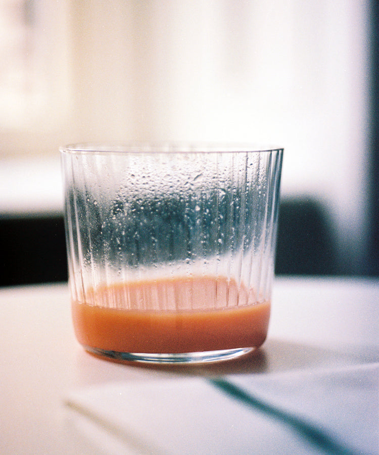 A clear, minimal Teo Ridged Tumbler from a set of 4 sits on a white surface, holding a small amount of orange liquid with condensation forming inside.