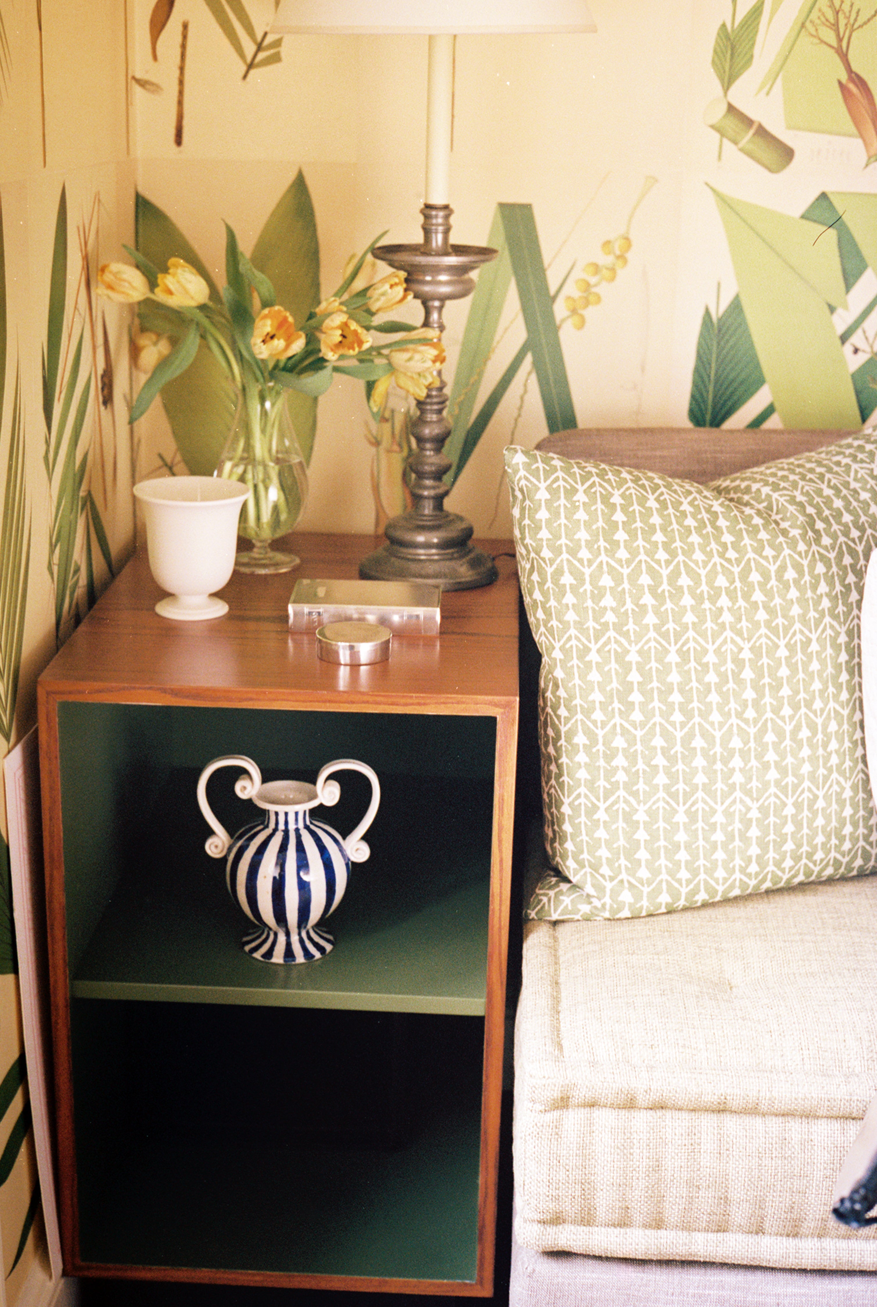A small wooden side table with a striped vase, lamp, white cup, yellow flowers in glass vase, and a green-patterned pillow on a beige sofa—perfect for displaying your Vintage Cigarette Box Book.