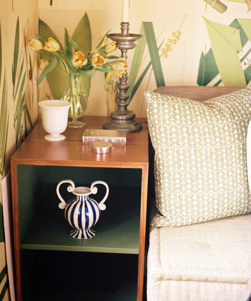A small wooden side table with a striped vase, lamp, white cup, yellow flowers in glass vase, and a green-patterned pillow on a beige sofa—perfect for displaying your Vintage Cigarette Box Book.