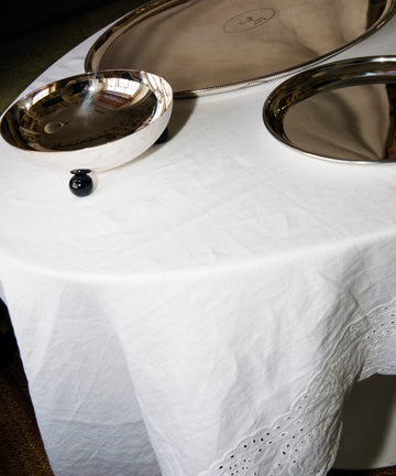 The Broderie Anglaise Linen Tablecloth, Medium, in white, covers the table and displays three silver serving trays of different shapes and sizes.