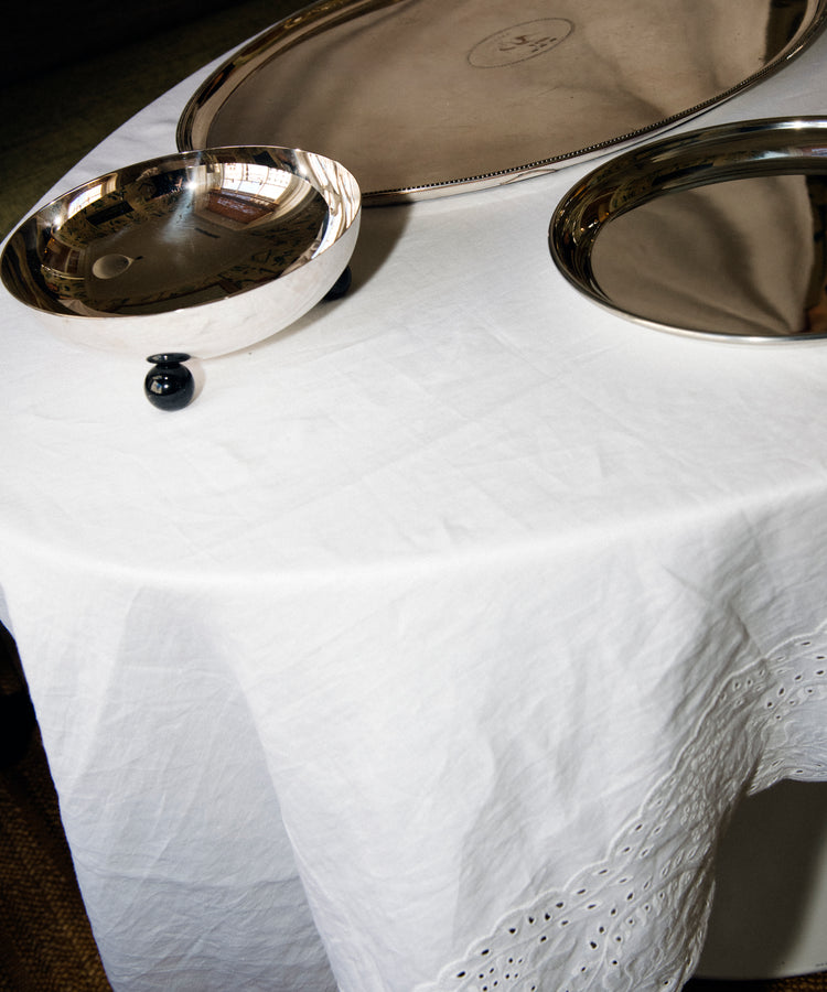 The Broderie Anglaise Linen Tablecloth, Medium, in white, covers the table and displays three silver serving trays of different shapes and sizes.