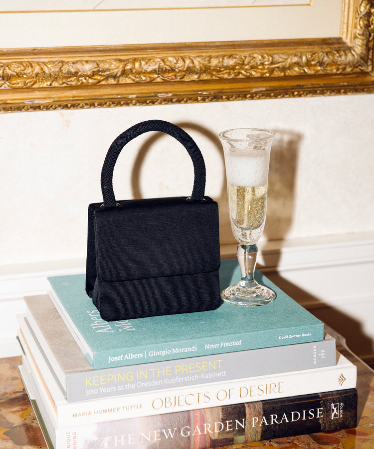 A Lauren Champagne Glass, filled with bubbly champagne, sits beside a small black handbag on a stack of books atop a marble surface, with framed artwork in the background.
