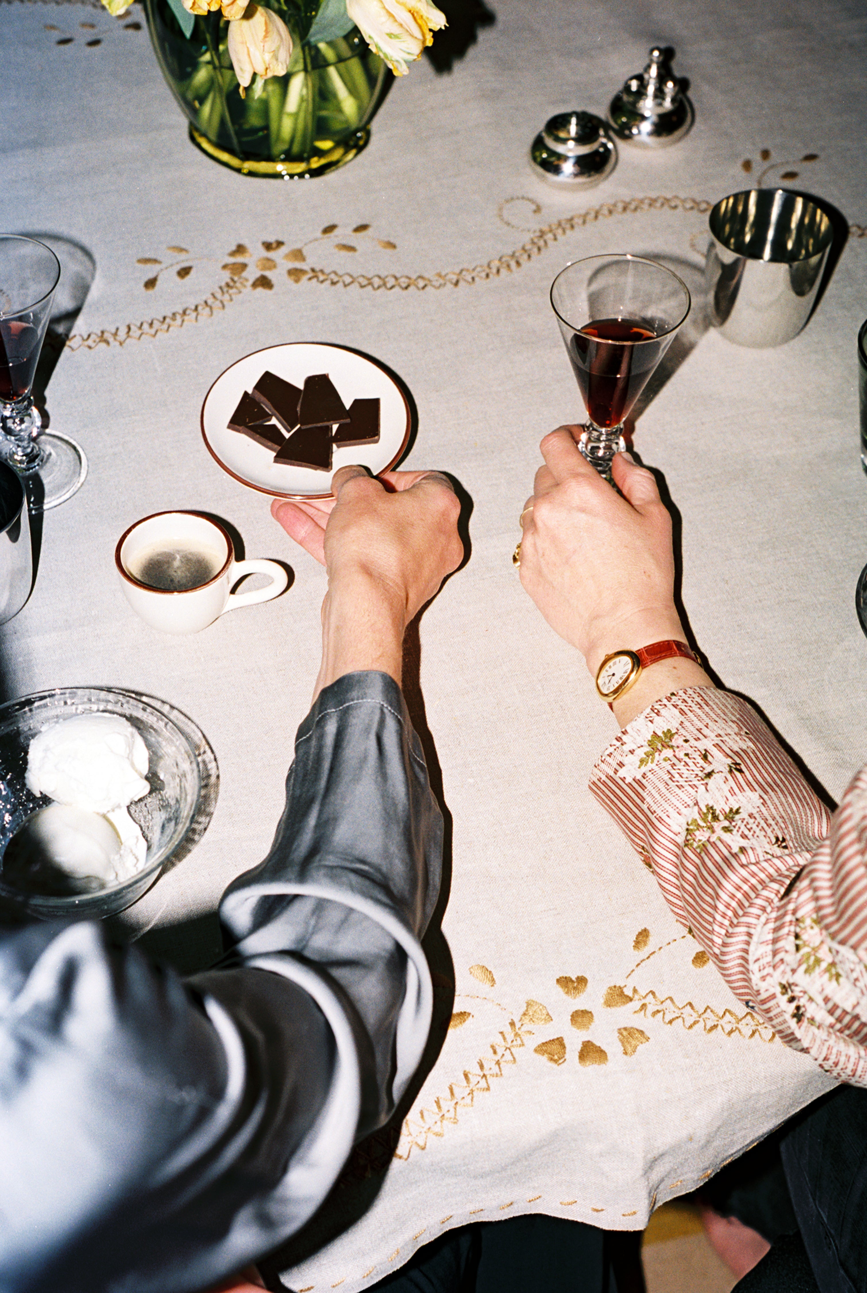 Two people sit at a table with wine glasses, coffee, chocolate pieces, and a bowl of whipped cream on the Zelia Tablecloth, featuring beautiful hand embroidery and traditional designs.