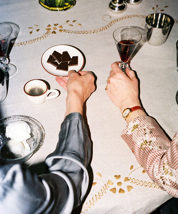 Two people sit at a table with wine glasses, coffee, chocolate pieces, and a bowl of whipped cream on the Zelia Tablecloth, featuring beautiful hand embroidery and traditional designs.