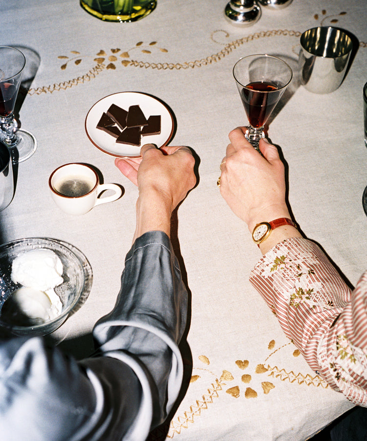 Two people sit at a table with wine glasses, coffee, chocolate pieces, and a bowl of whipped cream on the Zelia Tablecloth, featuring beautiful hand embroidery and traditional designs.
