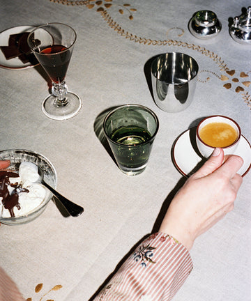 A person holds an espresso cup at a table featuring a Gem Glass, Set of 2, alongside a dessert bowl, glass of water, wine, and luxurious drinking glasses on a patterned tablecloth.