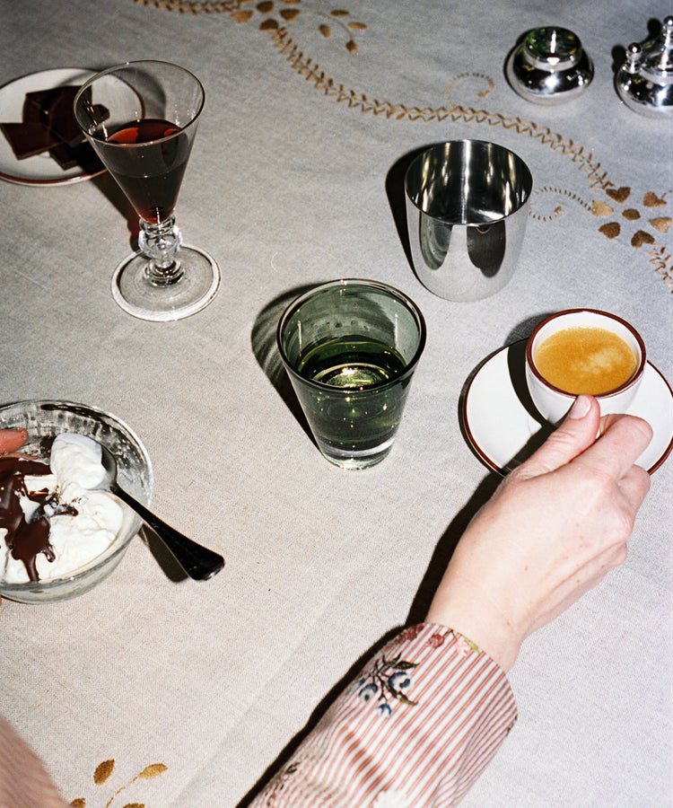 A person holds an espresso cup at a table featuring a Gem Glass, Set of 2, alongside a dessert bowl, glass of water, wine, and luxurious drinking glasses on a patterned tablecloth.