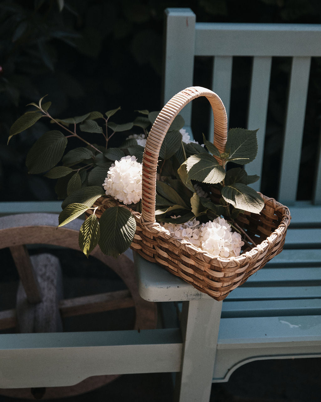 The Folk Fleur Hand Basket, filled with white flowers and green leaves, rests on the arm of a light blue wooden bench.