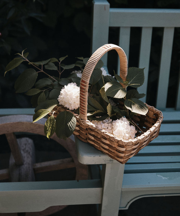 The Folk Fleur Hand Basket, filled with white flowers and green leaves, rests on the arm of a light blue wooden bench.