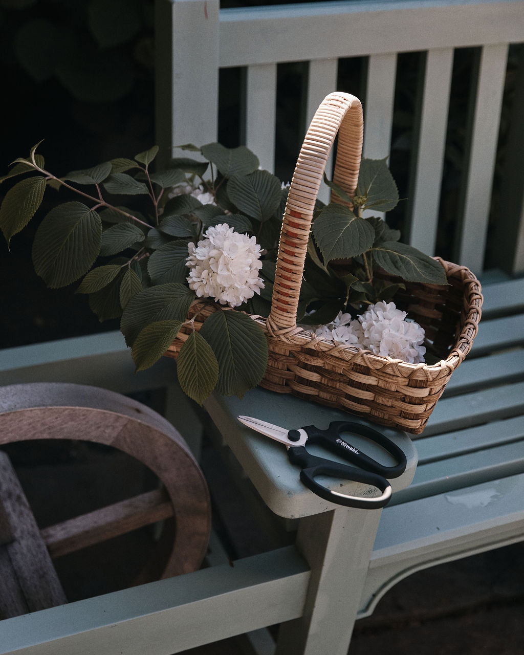 A woven basket brimming with white flowers and green leaves rests on a wooden bench beside a pair of Sakagen Flower Scissors and other gardening tools, ready for trimming stems.