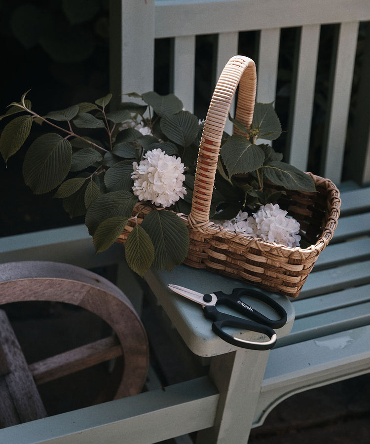 A woven basket brimming with white flowers and green leaves rests on a wooden bench beside a pair of Sakagen Flower Scissors and other gardening tools, ready for trimming stems.