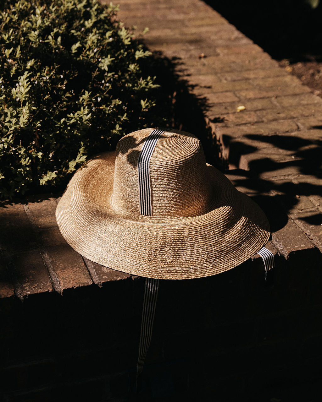 The Dahlia Hat, a natural straw piece with a relaxed silhouette and black-and-white striped ribbon, sits on a brick ledge beside a bush, partially in shadow.