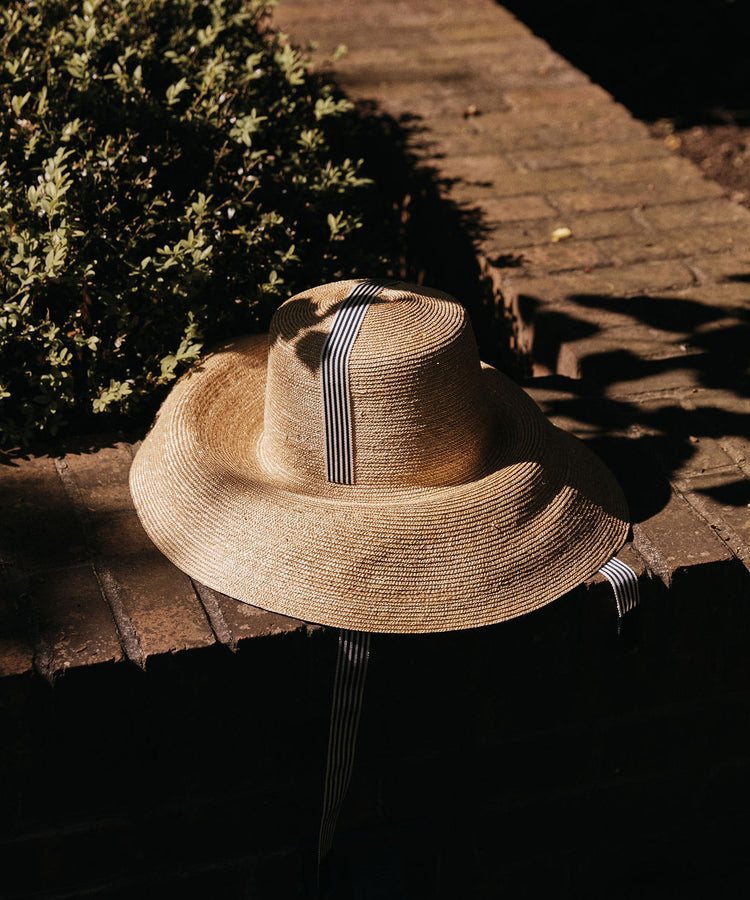 The Dahlia Hat, a natural straw piece with a relaxed silhouette and black-and-white striped ribbon, sits on a brick ledge beside a bush, partially in shadow.
