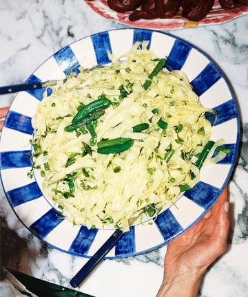 A person holds the Julius Serving Platter, a large blue and white dish with bold stripes, filled with tagliatelle pasta, peas, and fresh herbs on a marble countertop.