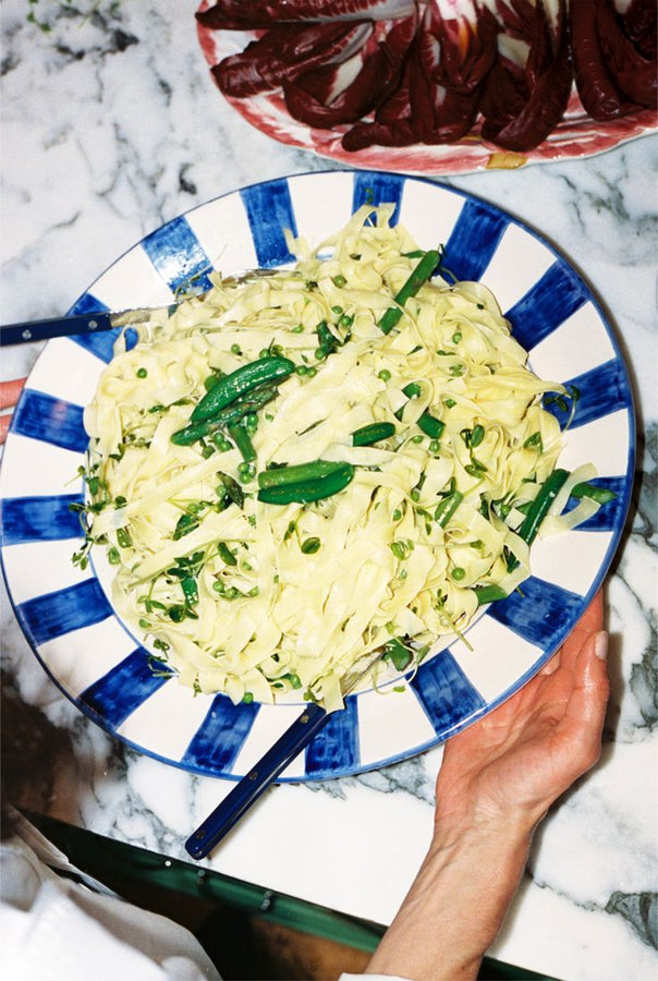 A person holds the Julius Serving Platter, a large blue and white dish with bold stripes, filled with tagliatelle pasta, peas, and fresh herbs on a marble countertop.