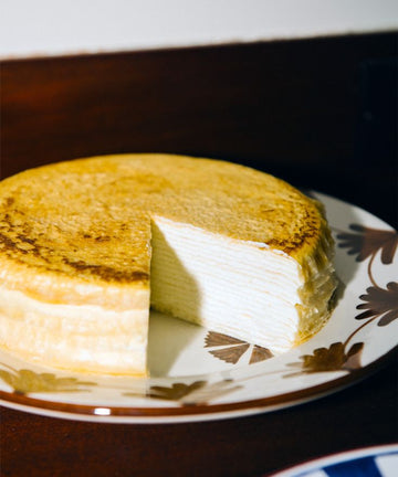 A round layered crepe cake with a slice removed sits atop a Catalina Small Plate from the set of 4, displayed on a wooden surface.
