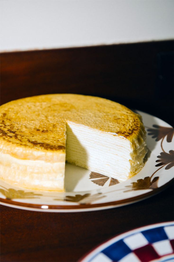 A round layered crepe cake with a slice removed sits atop a Catalina Small Plate from the set of 4, displayed on a wooden surface.