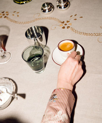 A person holding a Gio Espresso Set cup at a table set with a glass of water, coffee cup and saucer, silver containers, and an embroidered tablecloth.