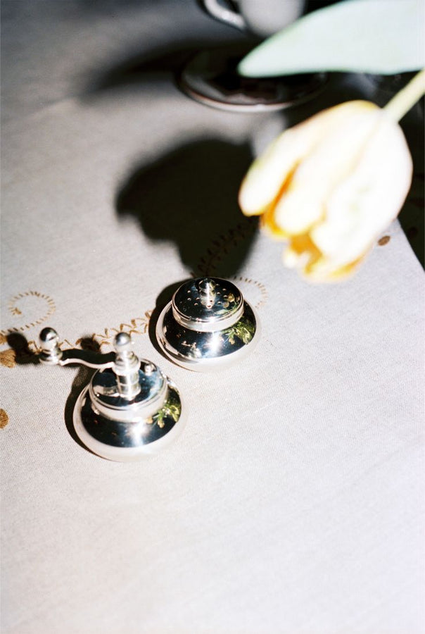 The Jules Pepper Mill and Salt Shaker rest on a light tablecloth, with a yellow tulip overhead casting a shadow—a subtle nod to refined Italian craftsmanship.