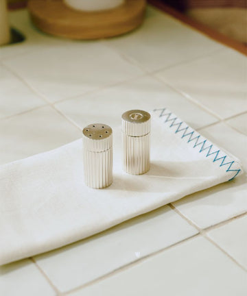 The Sophia Pepper Mill and Salt Shaker rest on a folded white cloth with blue stitching atop a tiled kitchen counter—a subtle nod to Italian craftsmanship.