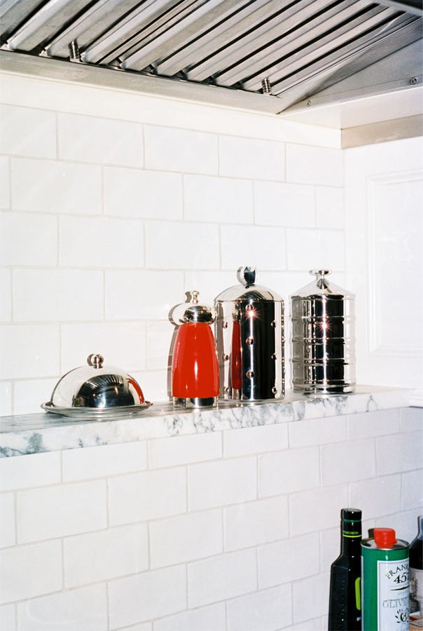 Four Kalisto Kitchen Box containers, including a red coffee pot and Italian-crafted stainless steel canisters, are lined up on a marble shelf against a white tiled backsplash. Bottles are partially visible below.