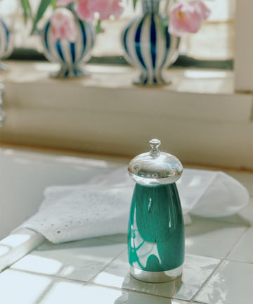 A teal and silver French press sits on a white tiled countertop, paired with the Giorgio Pepper Mill, while pink tulips in blue-striped vases brighten the background.