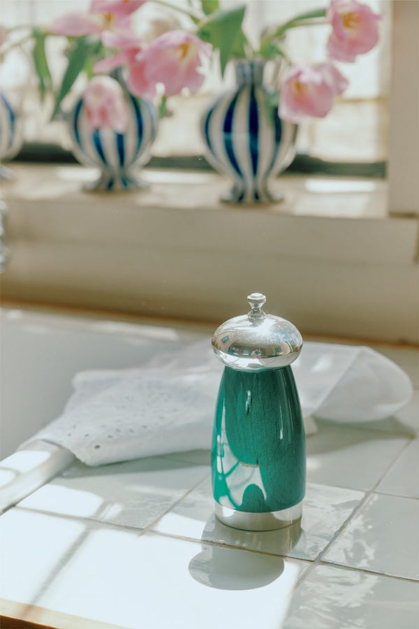 A teal and silver French press sits on a white tiled countertop, paired with the Giorgio Pepper Mill, while pink tulips in blue-striped vases brighten the background.