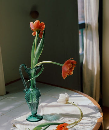 The Alba Vase, a hand-blown green piece, holds red and white tulips on a white tablecloth atop a round table near a window with sheer curtains, while some tulips are also arranged on the table.