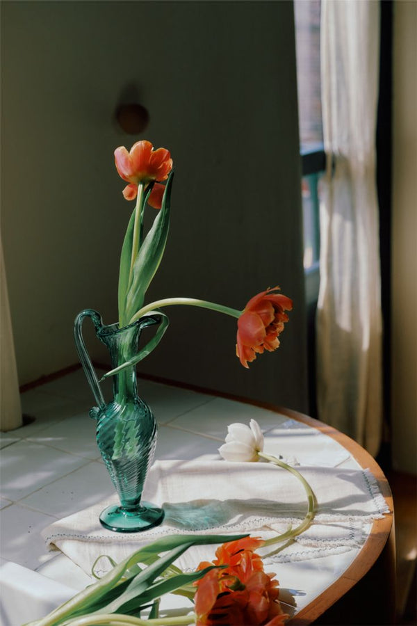 The Alba Vase, a hand-blown green piece, holds red and white tulips on a white tablecloth atop a round table near a window with sheer curtains, while some tulips are also arranged on the table.