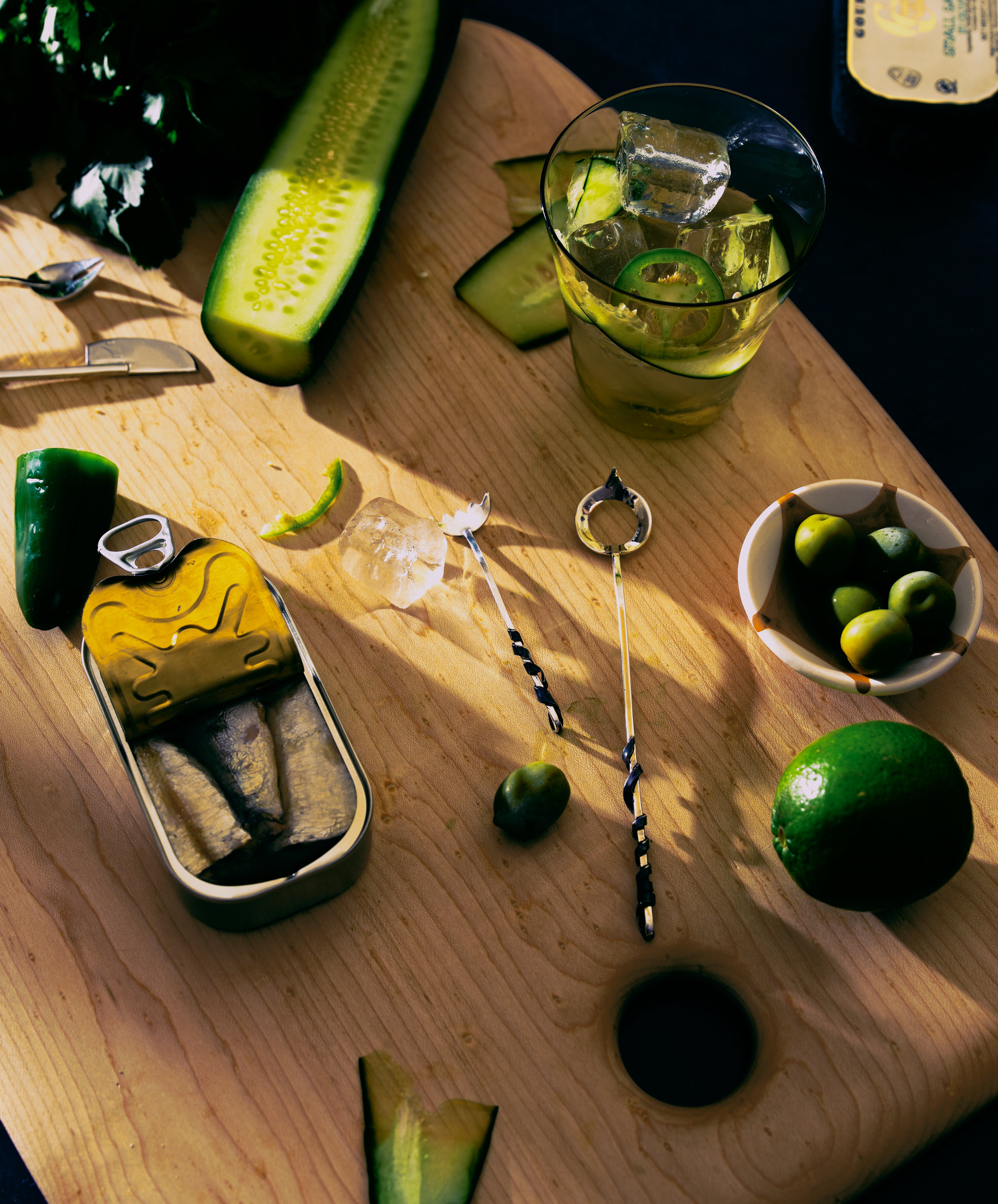 Olive spoon in situ on wooden board with olives, drink and tin of fish.