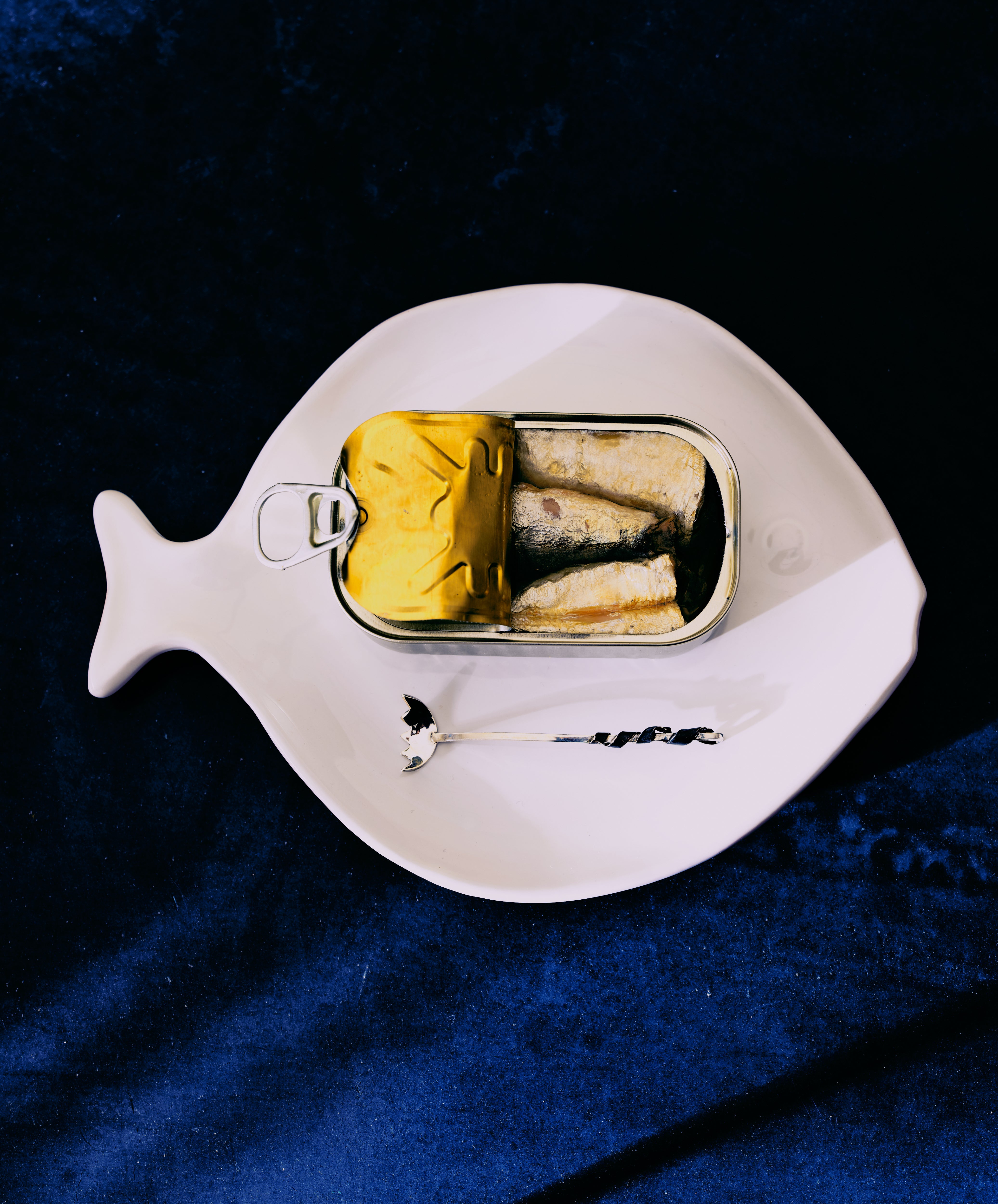 Silver fish fork in situ on white fish-shaped plate with  tinned fish. On blue fabric.