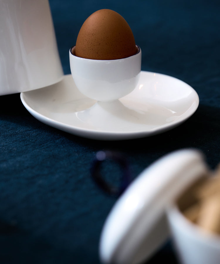 The Egg Cup and Lid holds a brown egg upright on a white plate, with a jar and container nearby on a dark blue tablecloth—ideal for your breakfast needs.