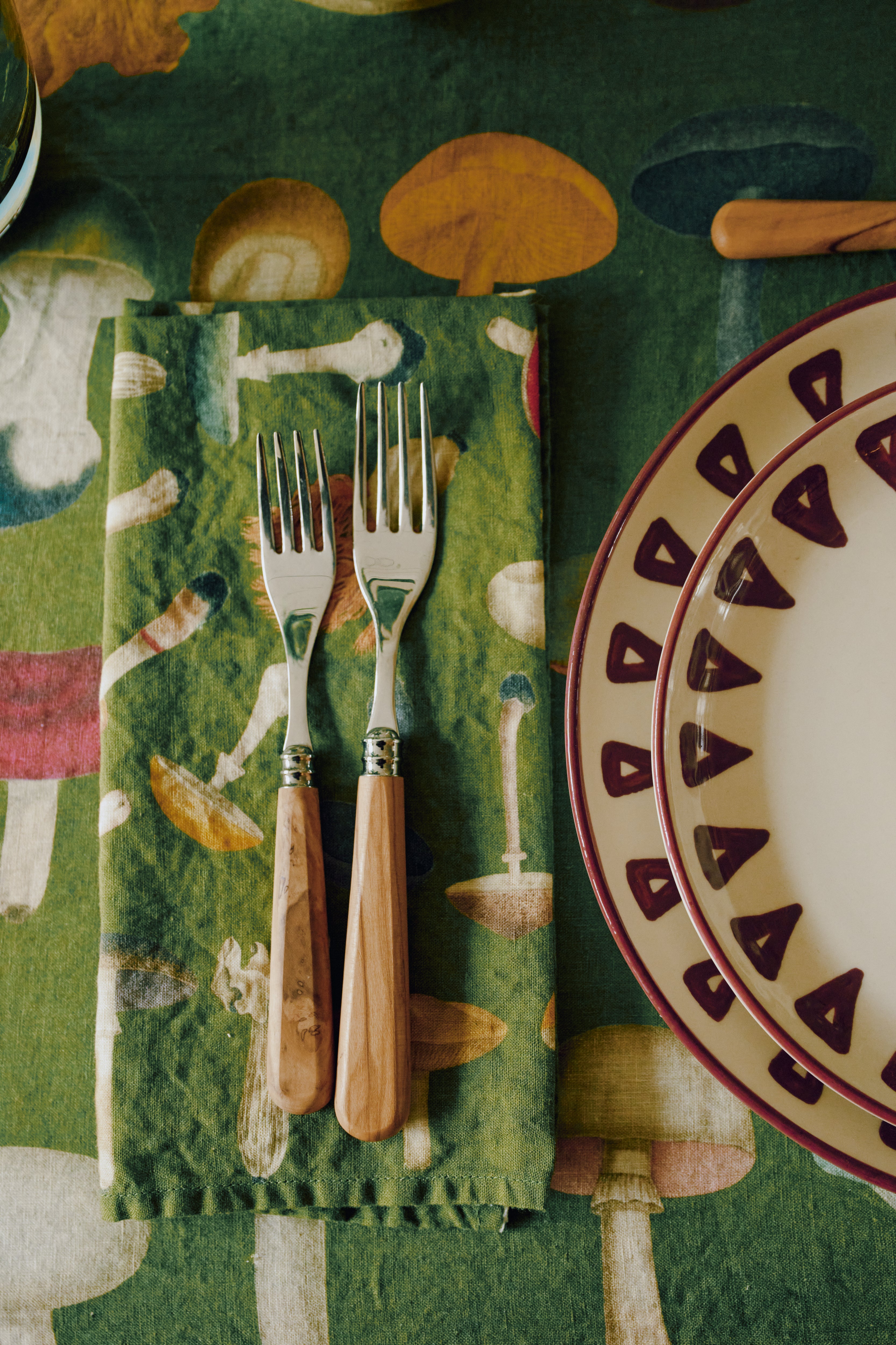 Two forks with wooden handles rest on a John Derian Fungi Napkin, Set of 6, their vintage mushroom motif standing out against the green fabric, next to a decorative plate with a maroon border atop a matching tablecloth.