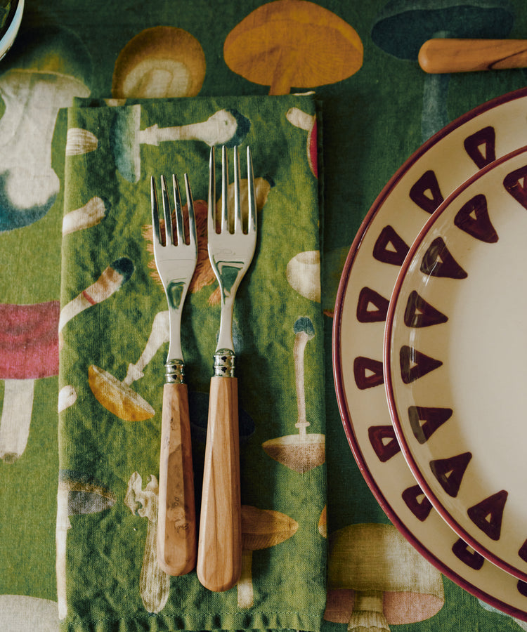 Two forks with wooden handles rest on a John Derian Fungi Napkin, Set of 6, their vintage mushroom motif standing out against the green fabric, next to a decorative plate with a maroon border atop a matching tablecloth.