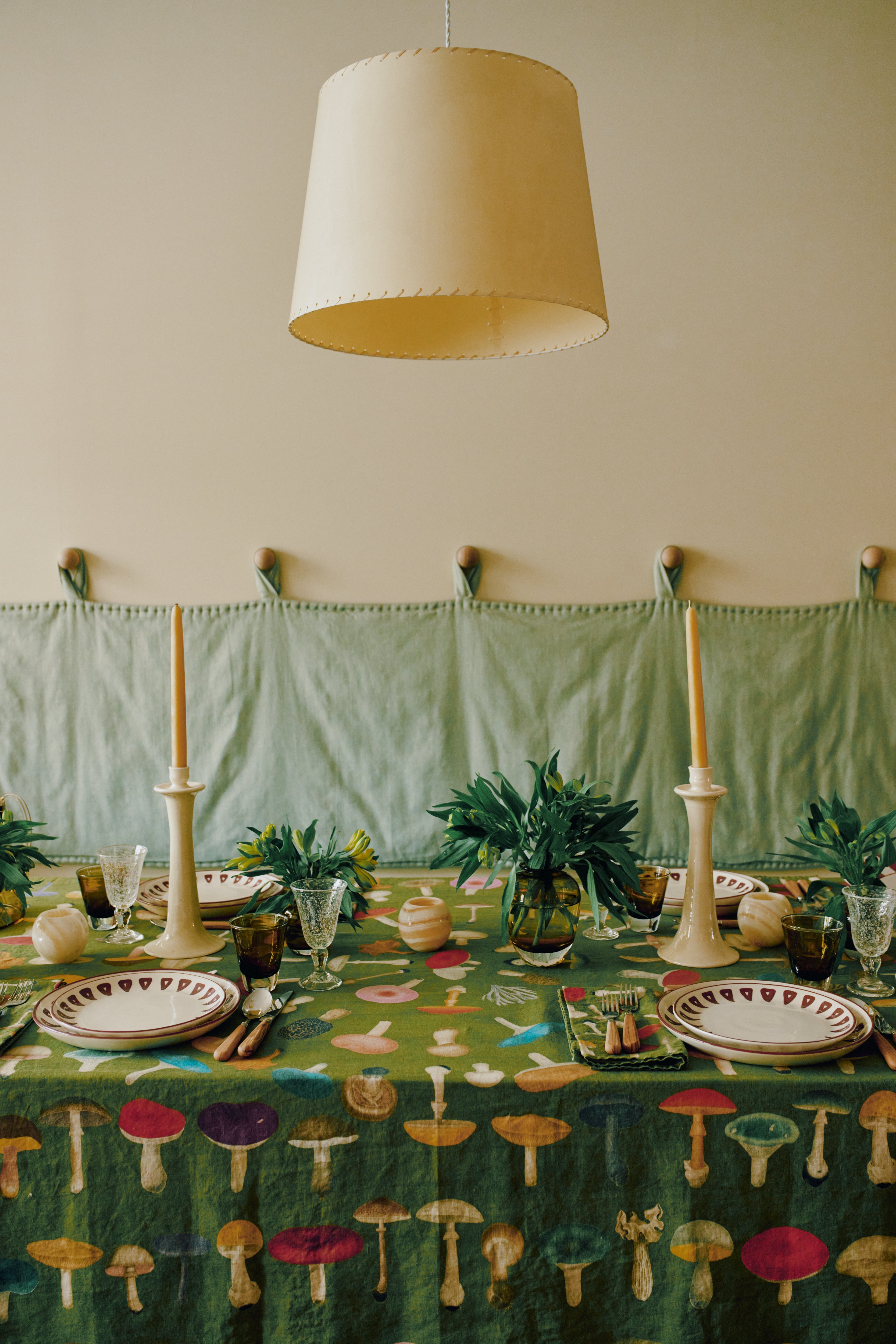 A dining table draped with the John Derian Fungi Tablecloth, complemented by two taper candles, plates, glassware, and green foliage vases, all arranged beneath a large beige lampshade.