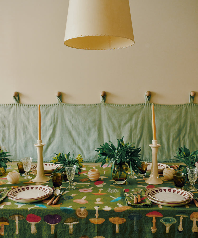 A dining table draped with the John Derian Fungi Tablecloth, complemented by two taper candles, plates, glassware, and green foliage vases, all arranged beneath a large beige lampshade.