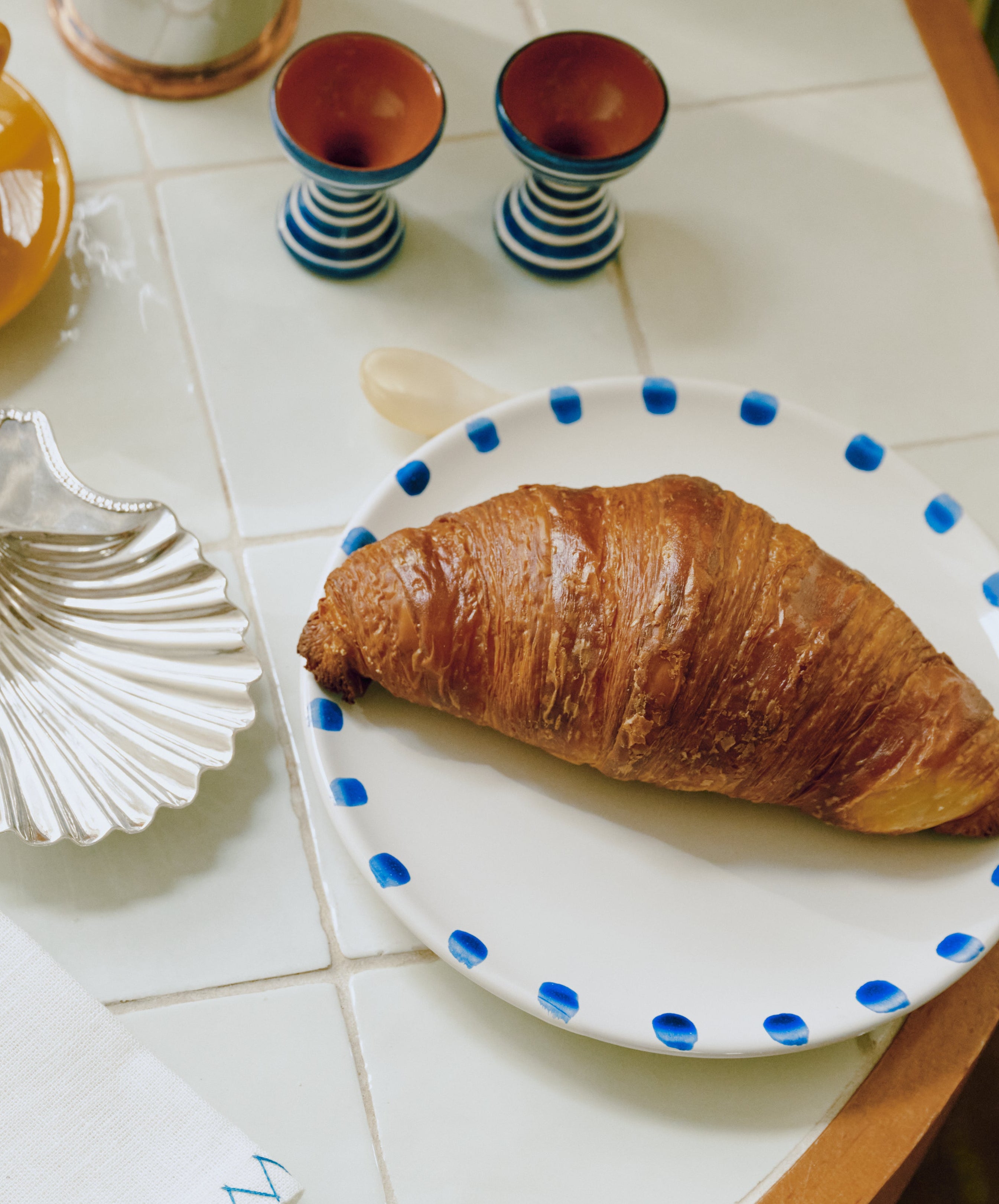 A flaky croissant rests on a Nina Small Plate, set of 4, with blue dots, placed on a tiled tabletop next to two small striped cups and a shell-shaped dish.