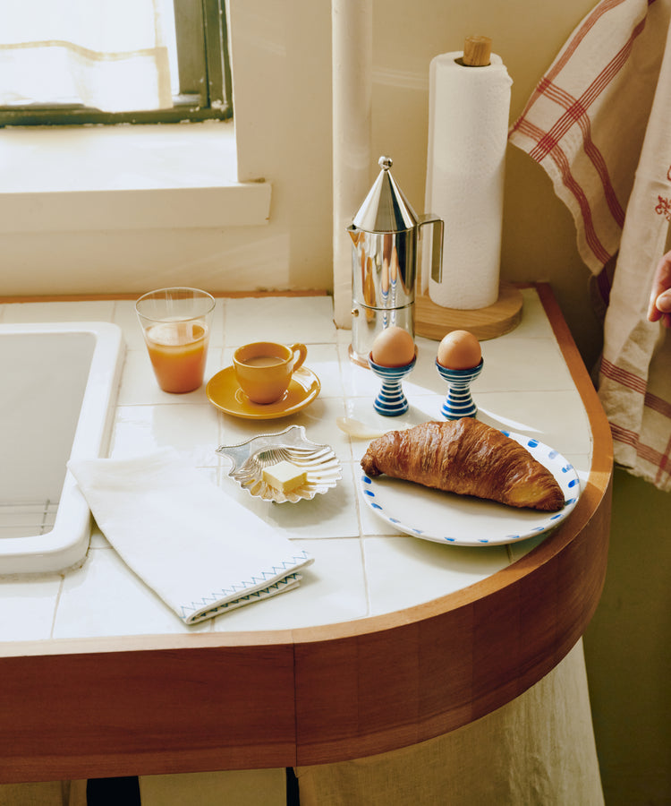 A croissant, two boiled eggs, a glass of orange juice, a napkin, and a cup of coffee made with the La Conica Espresso Coffee Maker are set on a tiled kitchen counter near a window.