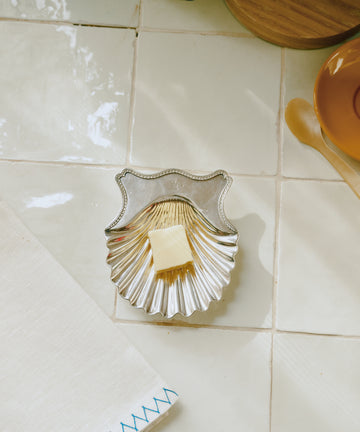 A small square of butter rests in a Vintage Silver Buttershells Pair dish—its shell-shaped design on a white tiled surface beside a napkin and plate.
