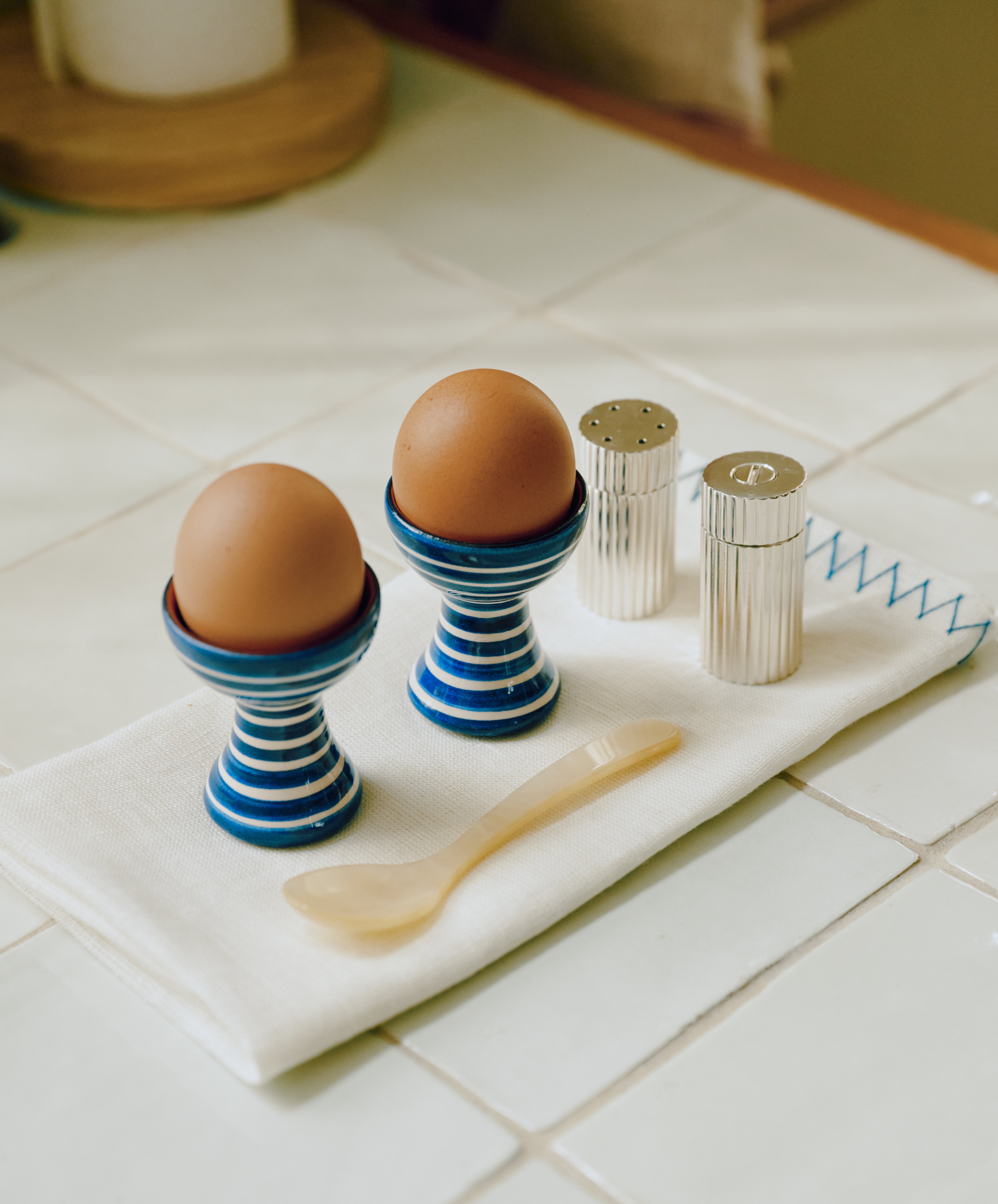 Two brown eggs in the Natalia Egg Cup, Set of 2—handmade with blue stripes—are paired with a beige spoon and silver salt and pepper shakers, all resting on a white cloth to create a charming breakfast set on the tiled kitchen counter.