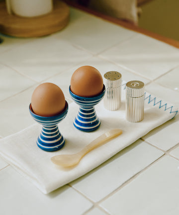 Two brown eggs in the Natalia Egg Cup, Set of 2—handmade with blue stripes—are paired with a beige spoon and silver salt and pepper shakers, all resting on a white cloth to create a charming breakfast set on the tiled kitchen counter.