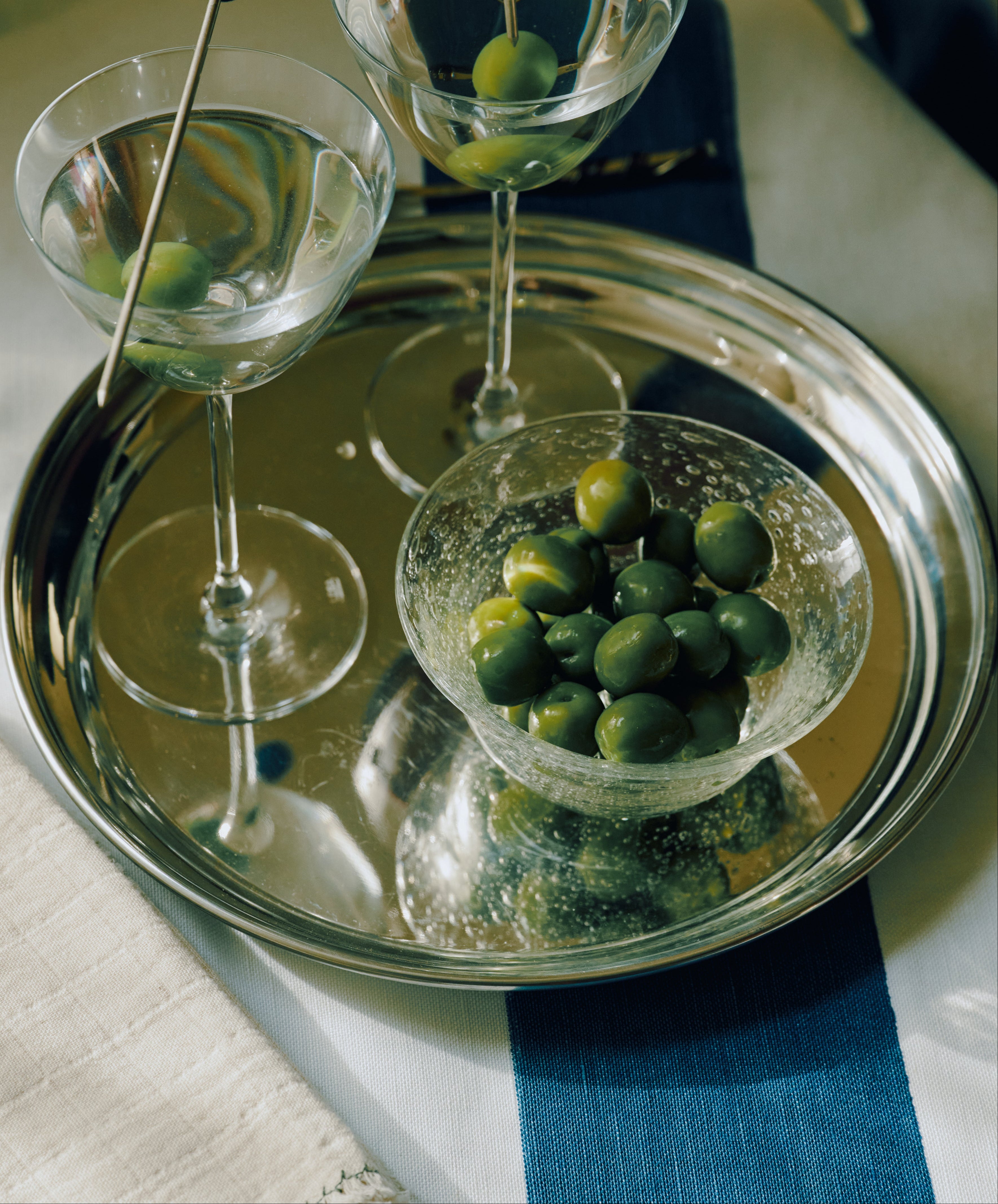 A silver serving tray holding two martini glasses with green olives, a small clear bowl of green olives, and a toothpick with a dark blue olive, set on a white and blue striped cloth.