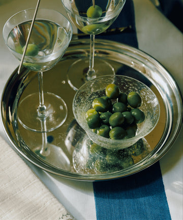 A silver serving tray holding two martini glasses with green olives, a small clear bowl of green olives, and a toothpick with a dark blue olive, set on a white and blue striped cloth.