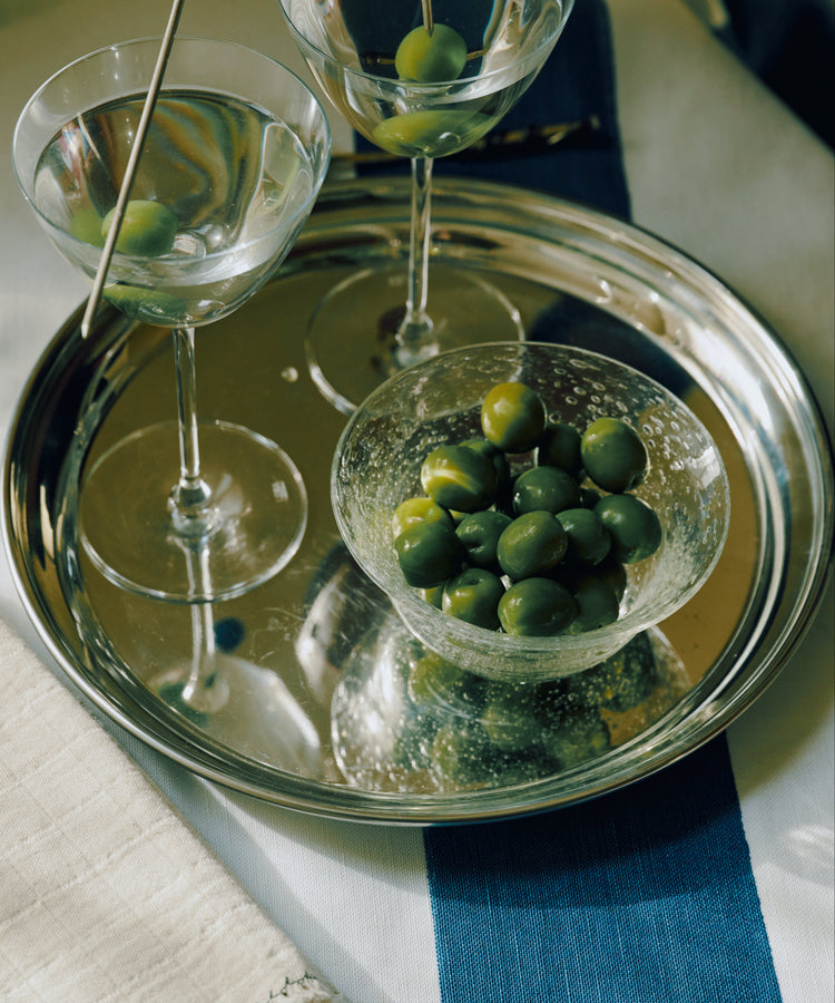 A silver serving tray holding two martini glasses with green olives, a small clear bowl of green olives, and a toothpick with a dark blue olive, set on a white and blue striped cloth.