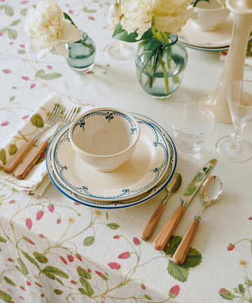 A neatly set dining table featuring the Arco Dinner Plate, Set of 4, paired with a floral tablecloth, wooden-handled cutlery, glasses, and vases of white flowers.