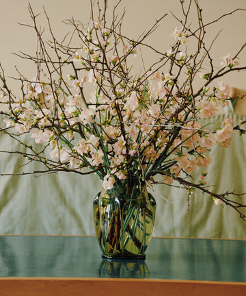 The Carlotta Vase, a handcrafted mouth-blown piece filled with long branches and pink blossoms, sits on a green tabletop and brings Bohemian artistry to a neutral background.
