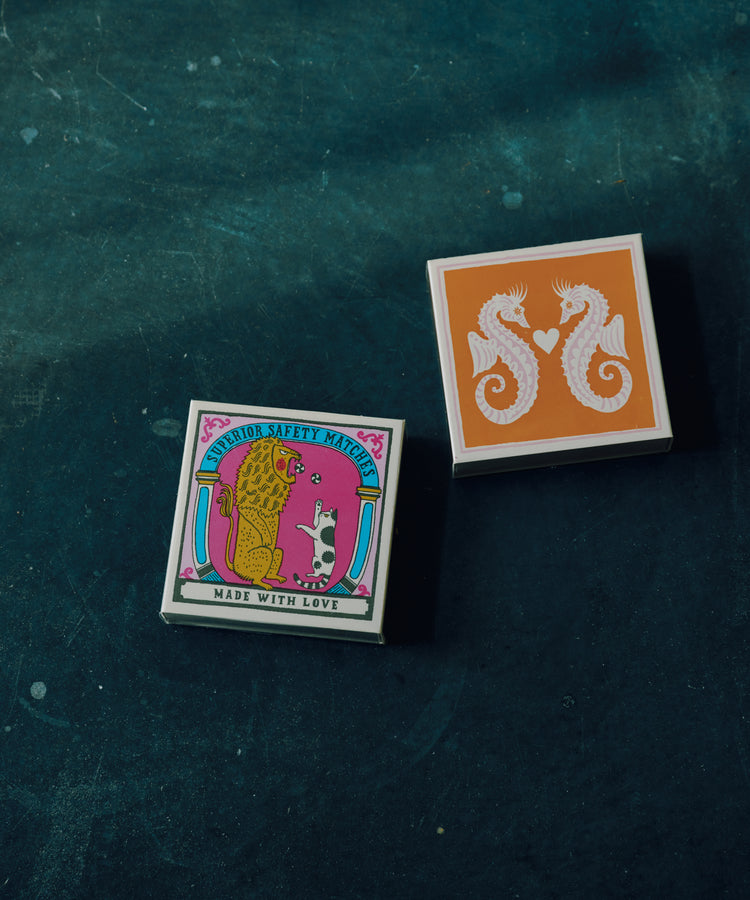 Two Luxury Matches matchboxes rest on a dark surface—one displays illustrated cats with “Made With Love,” the other shows seahorses with a heart—perfect for your partner in crime.