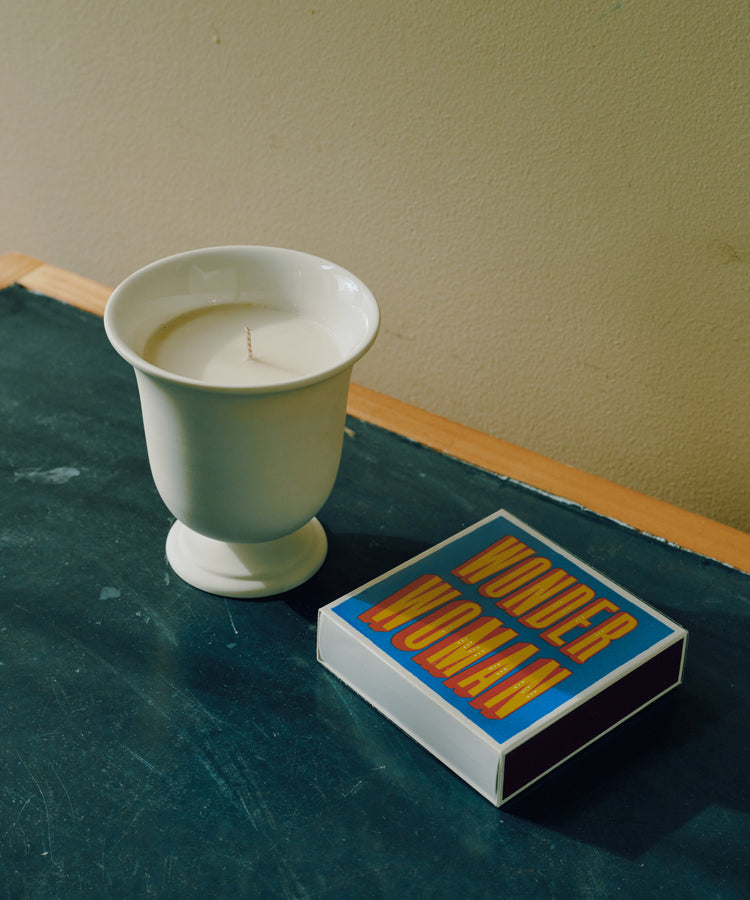 A white candle in a ceramic cup sits on a dark green surface beside a colorful “WONDER WOMAN” box and Luxury Matches, creating a cozy, charming vignette.