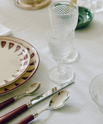 A set table with two patterned plates, three dark-handled silverware pieces, a green dish, and Celeste Red Wine Glasses on a white tablecloth.
